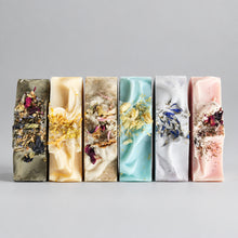 Six rectangular bars of artisanal soap arranged in a row on a light gray background. Each bar has a different pastel or earthy color and is topped with dried botanicals, including flowers and leaves.