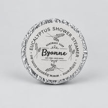 A round wrapped shower steamer from Byanne Handmade Soap, featuring a silver foil-wrapped edge and a light gray paper label with black text.