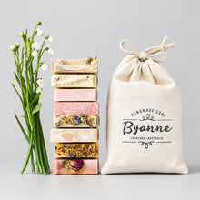 A set of handmade artisanal soaps in various colors and textures, arranged next to a natural beige cotton drawstring bag printed with 'Byanne Handmade Soap'