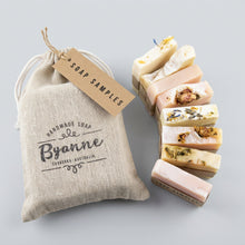 A set of handmade soap bars with visible botanical inclusions arranged next to a natural burlap drawstring bag printed with the brand name 'Byanne Handmade Soap' and a small 'SOAP SAMPLES' tag, 